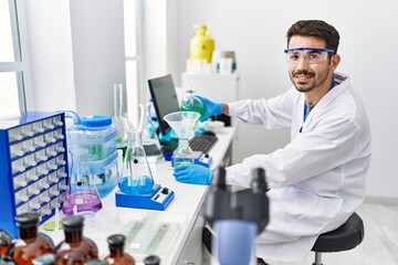 Fototapeta premium Young hispanic man wearing scientist uniform pouring liquid on test bottle at laboratory