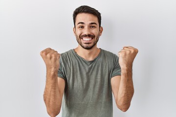 Fototapeta premium Young hispanic man with beard wearing casual t shirt over white background celebrating surprised and amazed for success with arms raised and open eyes. winner concept.