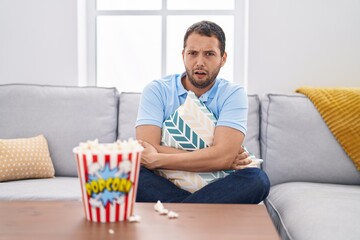 Hispanic man watching tv at home sitting on the sofa in shock face, looking skeptical and sarcastic, surprised with open mouth