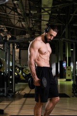 Fototapeta na wymiar A brutal muscular bearded man with a beautiful abs in headphones holds a dumbbell in the gym. Active lifestyle, sports