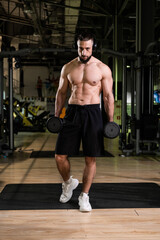 Fototapeta premium A brutal muscular bearded man with a beautiful abs in headphones holds a dumbbell in the gym. Active lifestyle, sports