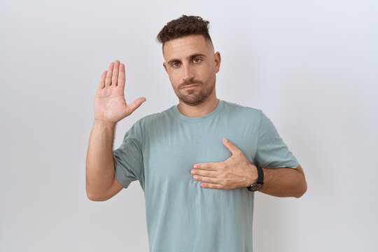 Hispanic man with beard standing over white background swearing with hand on chest and open palm, making a loyalty promise oath