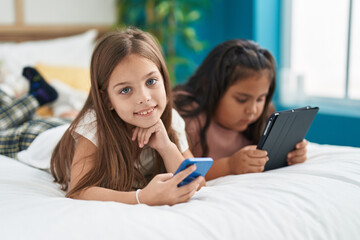 Two kids using smartphone and touchpad lying on bed at bedroom