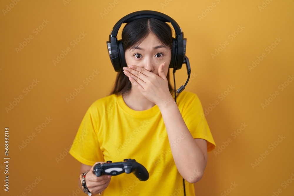 Sticker chinese young woman playing video game holding controller shocked covering mouth with hands for mist