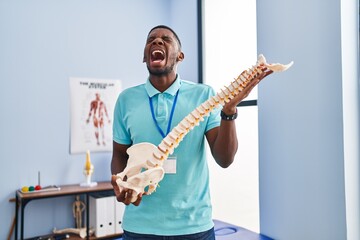 African american man holding anatomical model of spinal column angry and mad screaming frustrated...