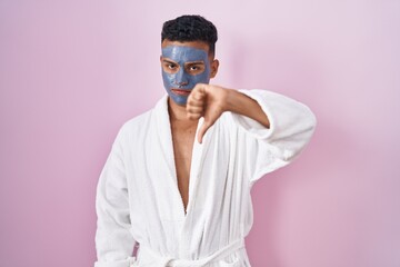 Young hispanic man wearing beauty face mask and bath robe looking unhappy and angry showing...