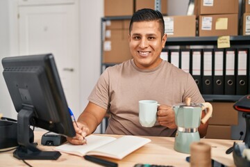 Young latin man ecommerce business worker drinking coffee writing on notebook at office