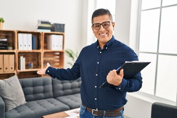 Fototapeta premium Young latin man business worker holding clipboard doing welcome gesture at office