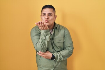 Fototapeta premium Hispanic young man standing over yellow background looking at the camera blowing a kiss with hand on air being lovely and sexy. love expression.