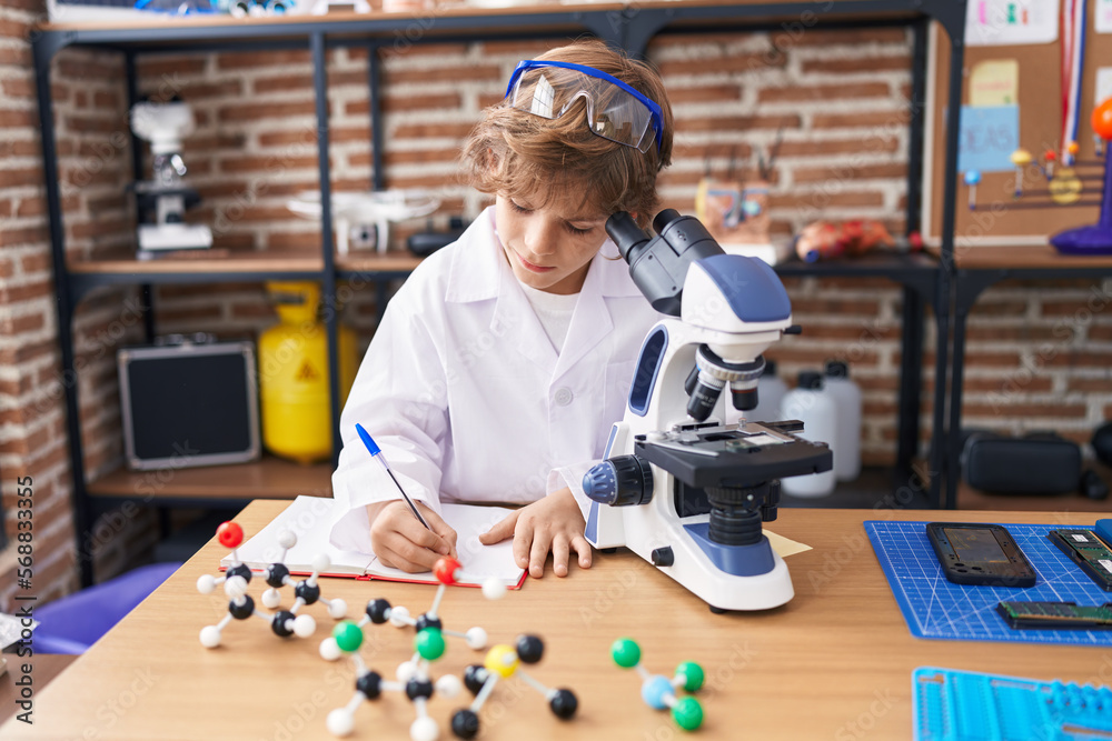 Poster adorable caucasian boy student using microscope writing on notebook at classroom