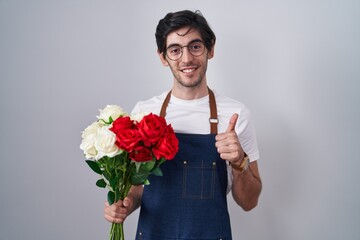 Young hispanic man holding bouquet of white and red roses doing happy thumbs up gesture with hand. approving expression looking at the camera showing success.