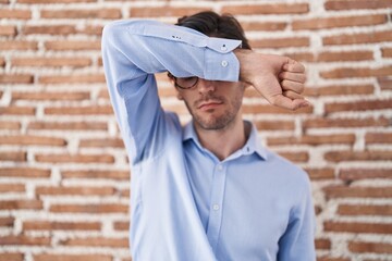Young hispanic man standing over brick wall background covering eyes with arm, looking serious and sad. sightless, hiding and rejection concept