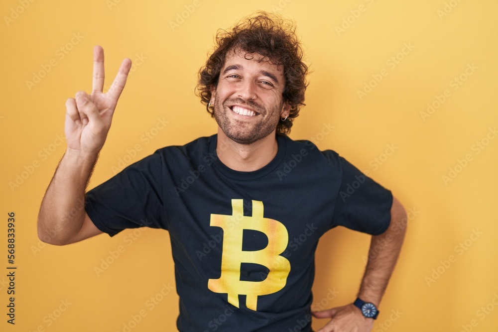 Canvas Prints Hispanic young man wearing bitcoin t shirt smiling looking to the camera showing fingers doing victory sign. number two.