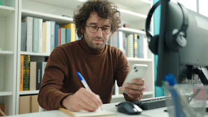 Fototapeta premium Young hispanic man student using smartphone writing on notebook at library university
