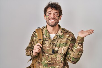 Hispanic young man wearing camouflage army uniform smiling cheerful presenting and pointing with palm of hand looking at the camera.