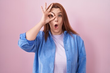 Young hispanic woman with red hair standing over pink background doing ok gesture shocked with surprised face, eye looking through fingers. unbelieving expression.