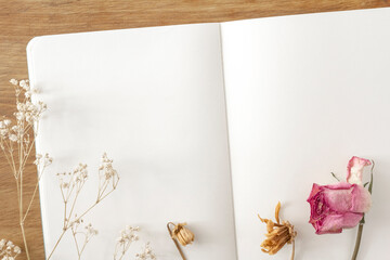 Blank notebook on a natural-looking wooden desk, and a lovely decorative frame composed of pink roses and other dried flowers on top of it.