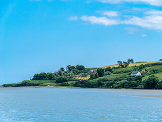 Beautiful clear sky over a calm water surface on a summer day. The picturesque green coast of Ireland. Several buildings on the hill. Seascape.