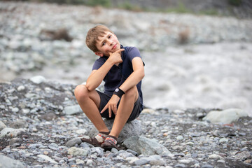 The boy is sitting on the bank of a mountain river.