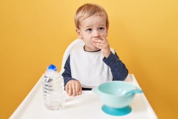 Adorable blond toddler sitting on highchair eating snack over isolated yellow background