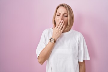 Young caucasian woman standing over pink background bored yawning tired covering mouth with hand....