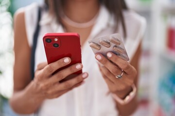 Young beautiful hispanic woman client using smartphone holding pills tablets at pharmacy