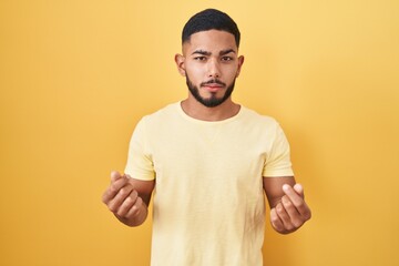 Young hispanic man standing over yellow background doing money gesture with hands, asking for salary payment, millionaire business