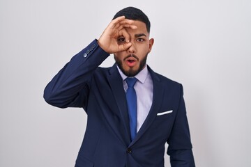 Young hispanic man wearing business suit and tie doing ok gesture shocked with surprised face, eye looking through fingers. unbelieving expression.