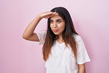 Young arab woman standing over pink background very happy and smiling looking far away with hand...