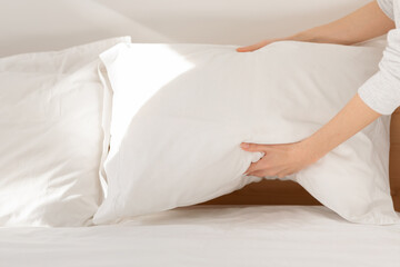Hands of caucasian millennial lady puts soft white pillow on comfortable bed, makes bed in bedroom