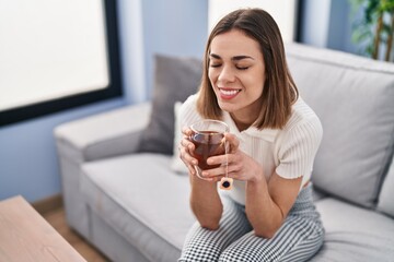 Young beautiful hispanic woman drinking tea sitting on sofa at home