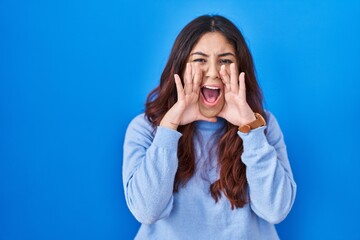 Hispanic young woman standing over blue background shouting angry out loud with hands over mouth