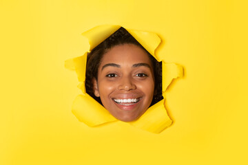 Excited african american woman posing in torn yellow paper background, looking at camera and smiling through hole
