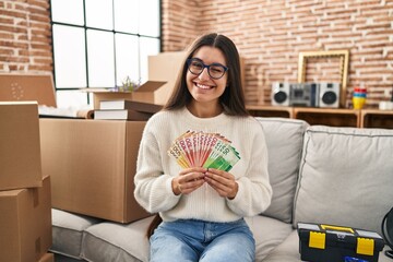 Young hispanic woman sitting on the sofa at new home holding money winking looking at the camera...