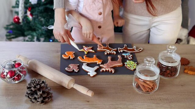 Happy little girl with her beautiful mom coloring Christmas gingerbread cookies at home. Christmas decoration in the kitchen. Fir tree with fairy lights. The concept of the New Year and Christmas. Hol