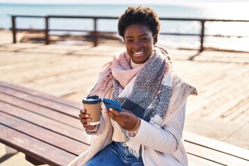 Obraz premium African american woman using smartphone drinking coffee sitting on bench at seaside