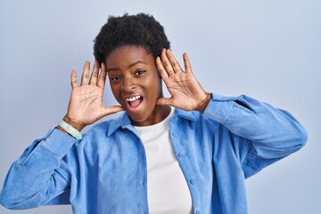African american woman standing over blue background smiling cheerful playing peek a boo with hands showing face. surprised and exited
