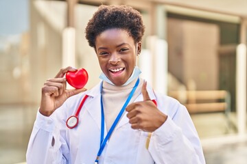 African american woman wearing doctor uniform holding heart smiling happy and positive, thumb up...