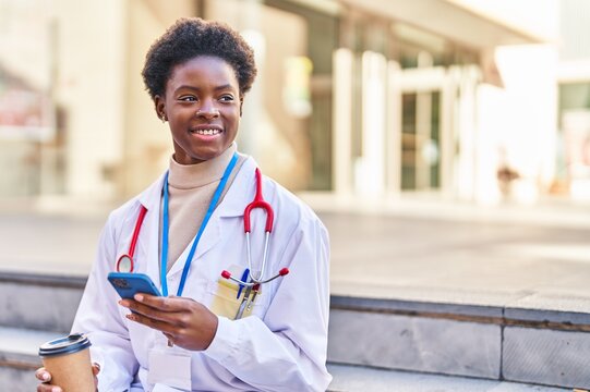 African american woman wearing doctor uniform using smartphone drinking coffee at street