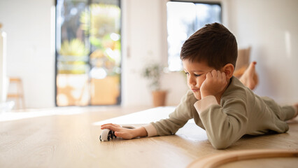 Busy serious bored caucasian little kid lying on floor, playing with toys cars or train, thinking...