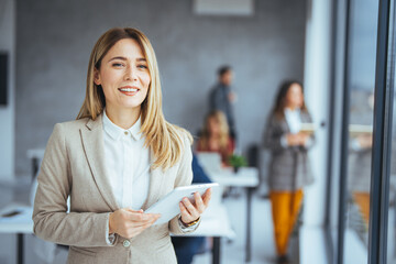 Portrait of happy blond businesswoman using digital tablet in agency. Successful business woman in casual clothing working on tablet. Mixed race young woman looking at camera in creative office.