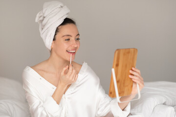 Glad caucasian millennial lady in towel, applies gloss on lips, looks in mirror, enjoys good morning in bedroom