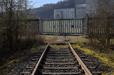 Würgassen, Germany - 03/23/2020: Old train tracks run towards a massive metal gate. There are bushes on the left and right. In the background are buildings from the nuclear power plant.