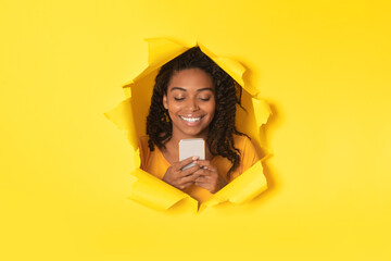 Happy african american lady using cellphone and browsing internet, posing in torn hole in yellow paper background