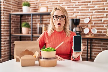 Blonde woman eating take away food showing smartphone screen scared and amazed with open mouth for surprise, disbelief face