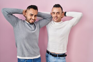 Homosexual couple standing over pink background relaxing and stretching, arms and hands behind head...