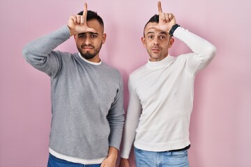 Homosexual couple standing over pink background making fun of people with fingers on forehead doing...