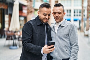 Two men couple smiling confident using smartphone at coffee shop terrace