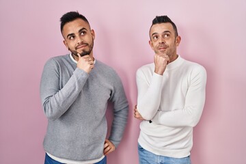 Homosexual couple standing over pink background with hand on chin thinking about question, pensive expression. smiling with thoughtful face. doubt concept.