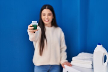 Young beautiful hispanic woman smiling confident holding detergent at laundry room